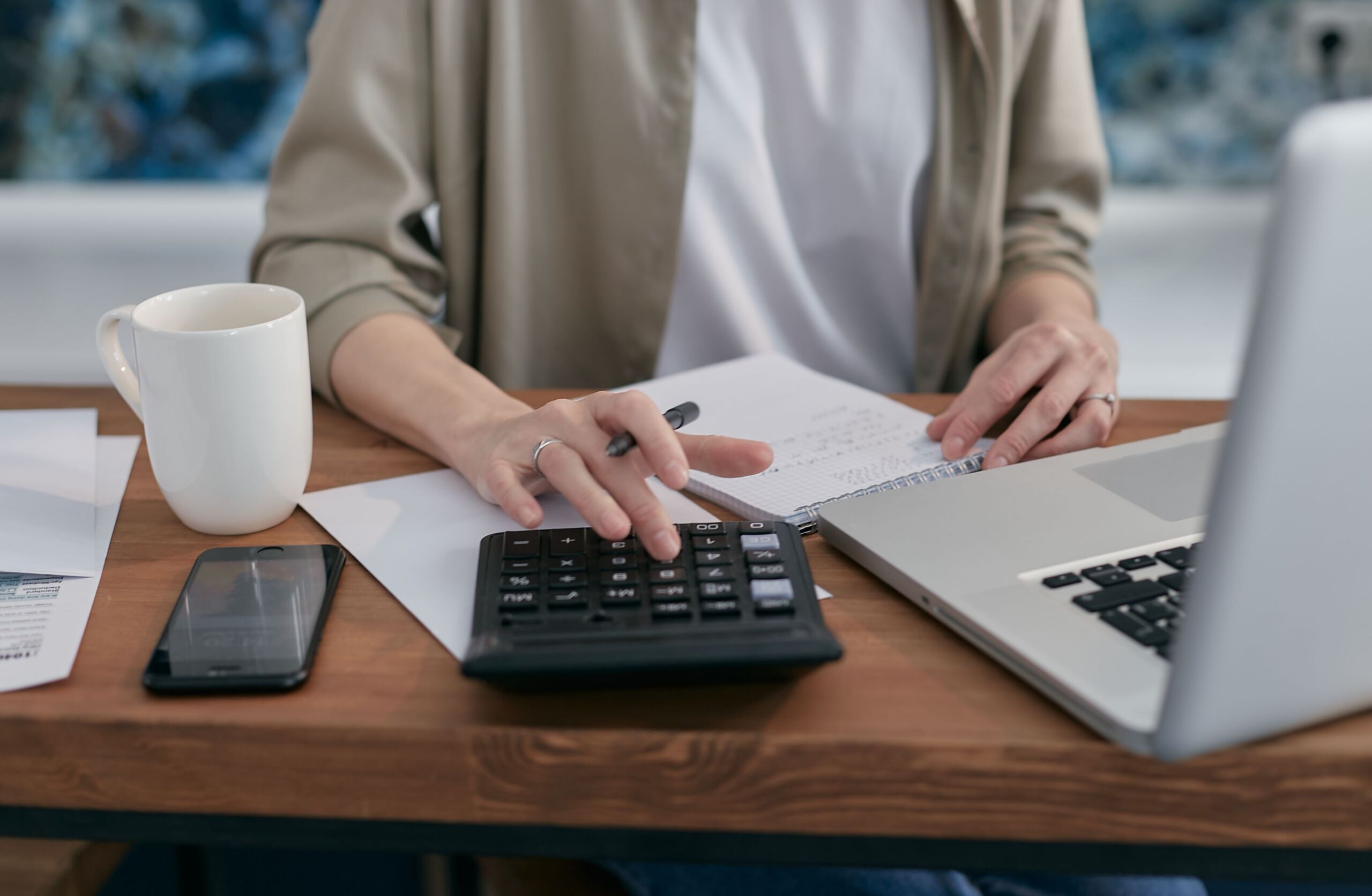 a person sitting at a wooden table with a notepad and laptop while using a calculator